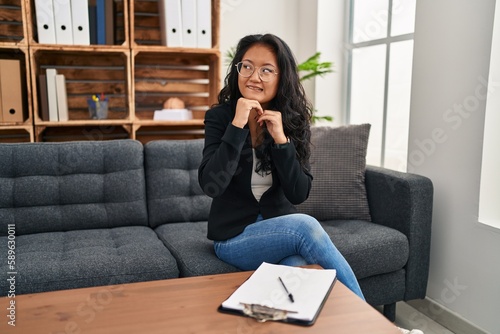 Young asian woman at consultation office laughing nervous and excited with hands on chin looking to the side