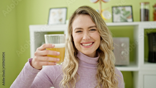 Young blonde woman holding glass of orange juice sitting on table at dinning room