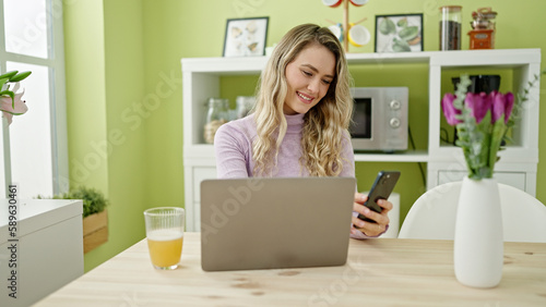 Young blonde woman using laptop and smartphone at dinning room