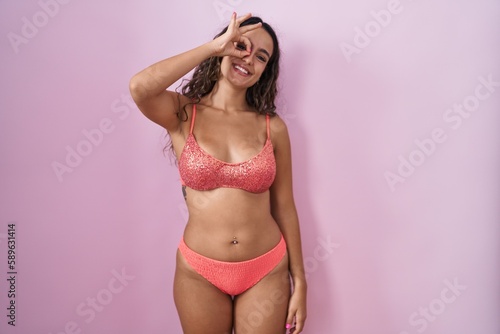 Young hispanic woman wearing lingerie over pink background doing ok gesture with hand smiling, eye looking through fingers with happy face.