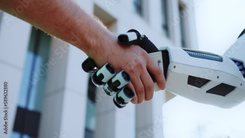 Horizontal view of male hand shaking robot hand outdoors. Blurred background of white building. Modern technologies. Connection of human and artificial intelligence. Future concept