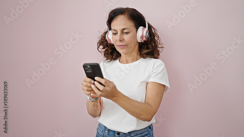 Middle age hispanic woman using smartphone wearing headphones over isolated pink background