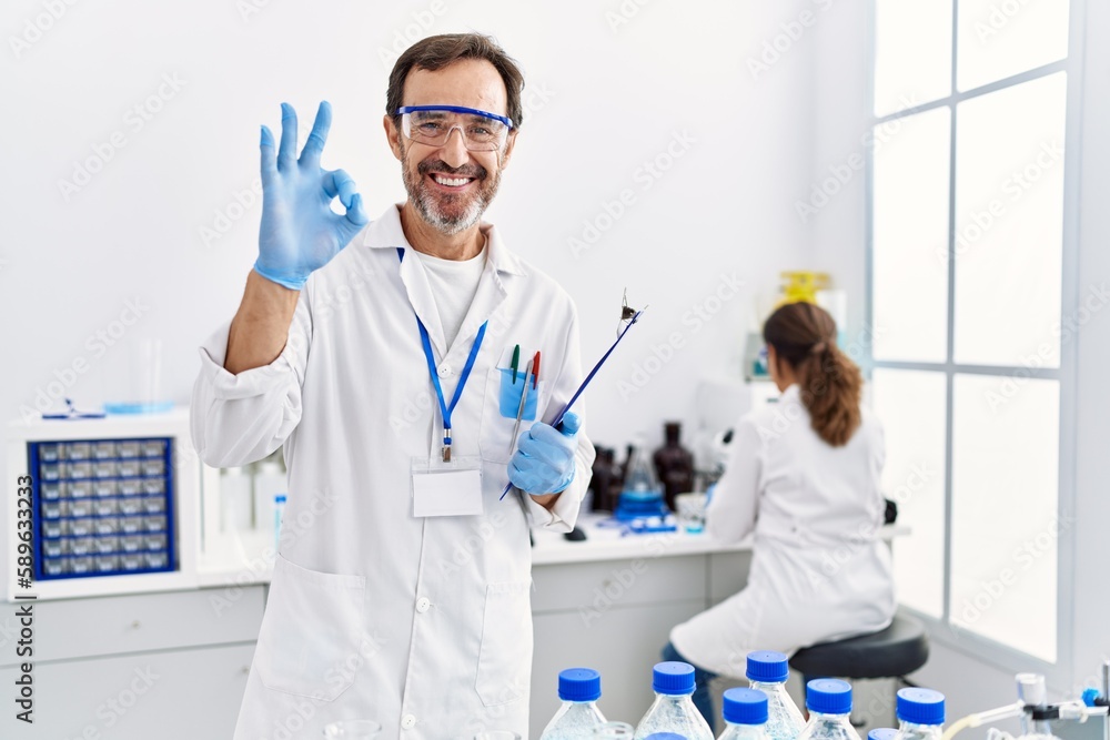 Middle age man working at scientist laboratory doing ok sign with fingers, smiling friendly gesturing excellent symbol