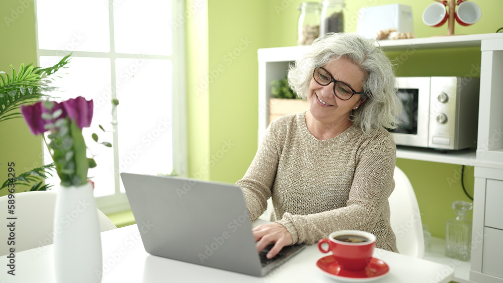 Middle age woman with grey hair using laptop sitting on table at home Middle age woman with grey hair using laptop sitting on table at home