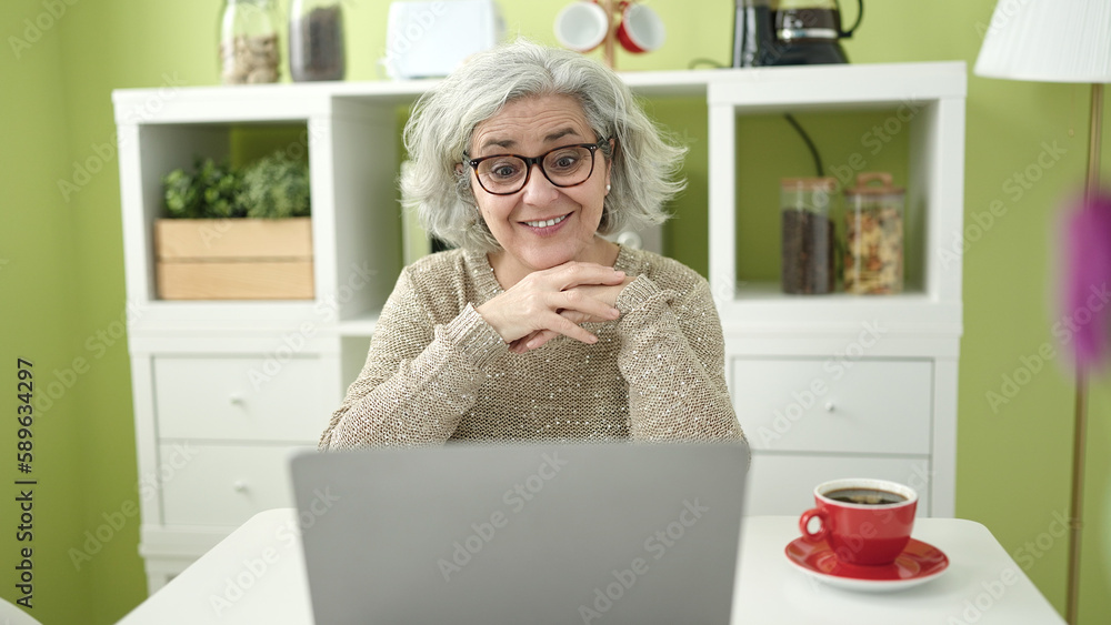 Middle age woman with grey hair having video call sitting on table at home Middle age woman with grey hair having video call sitting on table at home