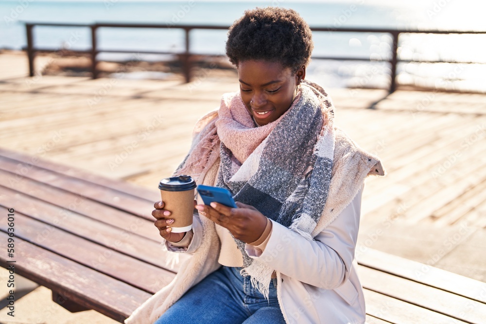 Obraz premium African american woman using smartphone drinking coffee sitting on bench at seaside
