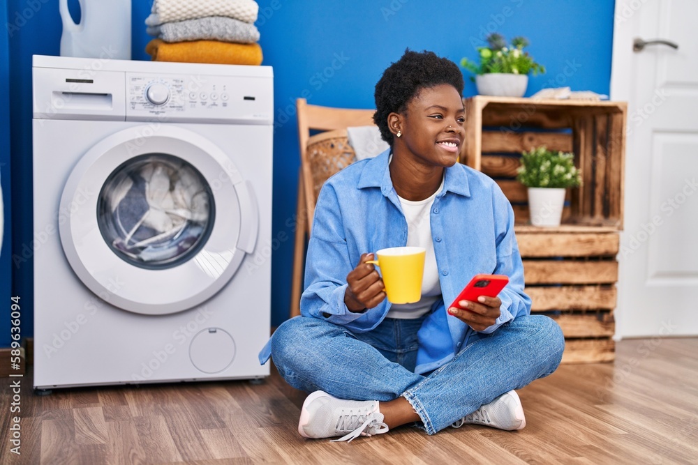 African american woman using smartphone and drinking coffee waiting for washing machine at laundry room African american woman using smartphone and drinking coffee waiting for washing machine at laundry room