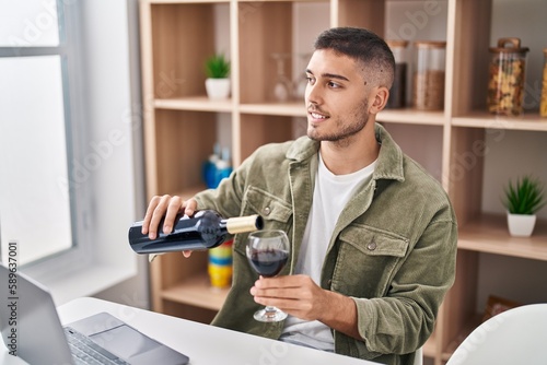Young hispanic man using laptop pouring wine on glass at home