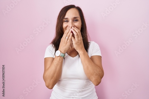 Middle age brunette woman standing over pink background laughing and embarrassed giggle covering mouth with hands, gossip and scandal concept