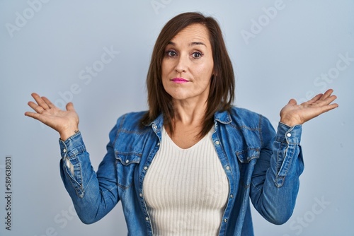 Middle age brunette woman wearing casual denim jacket over isolated background clueless and confused expression with arms and hands raised. doubt concept.