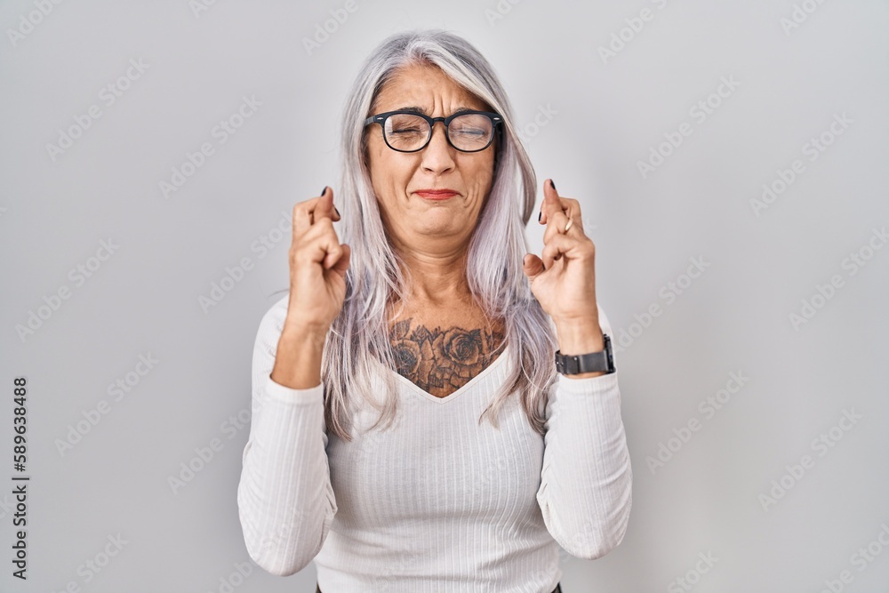 Middle age woman with grey hair standing over white background gesturing finger crossed smiling with hope and eyes closed. luck and superstitious concept. Middle age woman with grey hair standing over white background gesturing finger crossed smiling with hope and eyes closed. luck and superstitious concept.
