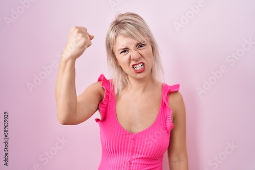 Young caucasian woman standing over pink background angry and mad raising fist frustrated and furious while shouting with anger. rage and aggressive concept.