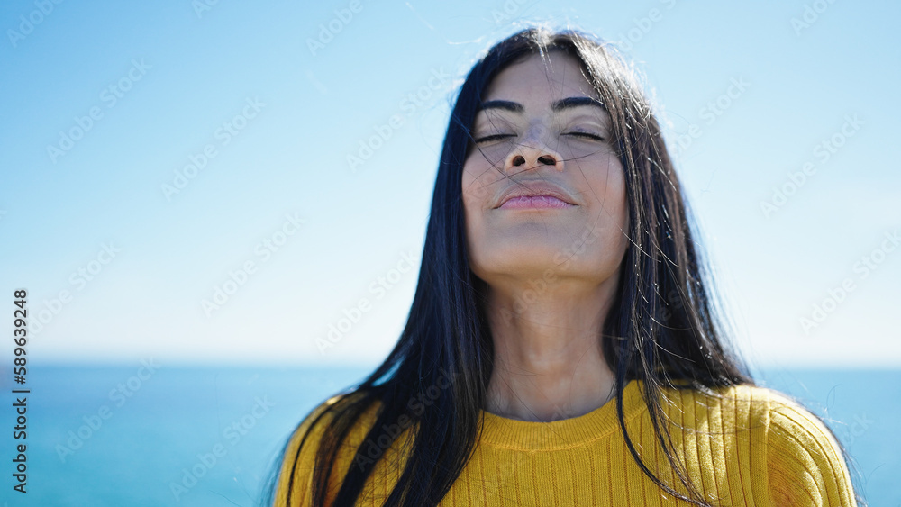 Young beautiful hispanic woman breathing with closed eyes at seaside