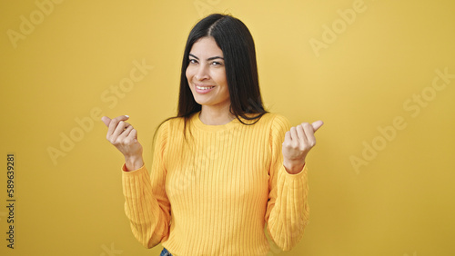 Young beautiful hispanic woman smiling confident doing money gesture over isolated yellow background