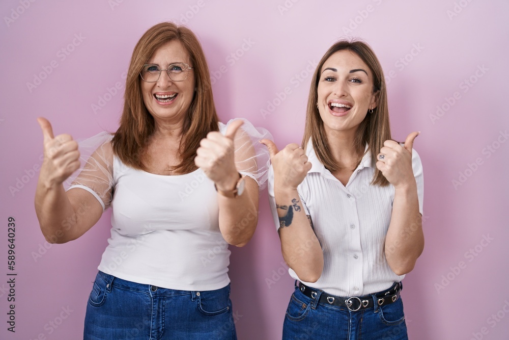 Hispanic mother and daughter wearing casual white t shirt over pink background success sign doing positive gesture with hand, thumbs up smiling and happy. cheerful expression and winner gesture. Hispanic mother and daughter wearing casual white t shirt over pink background success sign doing positive gesture with hand, thumbs up smiling and happy. cheerful expression and winner gesture.