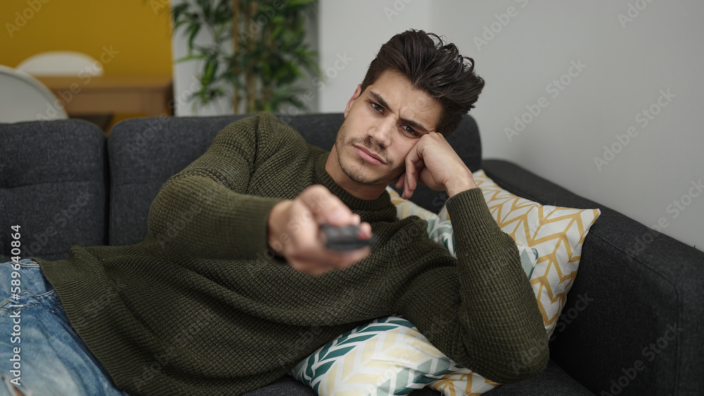 Young hispanic man watching tv lying on sofa with boring expression at home Young hispanic man watching tv lying on sofa with boring expression at home
