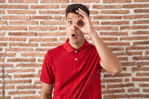 Young hispanic man standing over bricks wall doing ok gesture shocked with surprised face, eye looking through fingers. unbelieving expression.