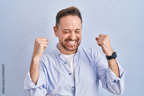 Middle age caucasian man standing over blue background excited for success with arms raised and eyes closed celebrating victory smiling. winner concept.