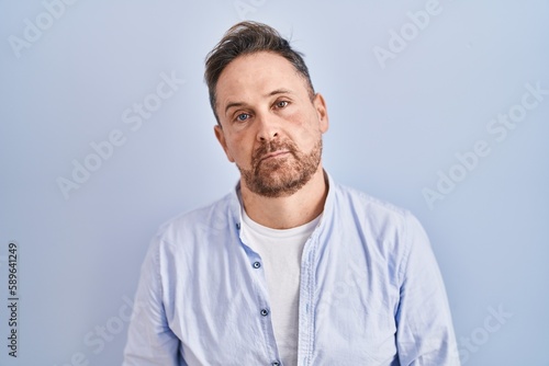 Middle age caucasian man standing over blue background looking sleepy and tired, exhausted for fatigue and hangover, lazy eyes in the morning.