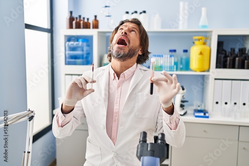 Handsome middle age man holding blood sample at biology laboratory angry and mad screaming frustrated and furious, shouting with anger looking up.