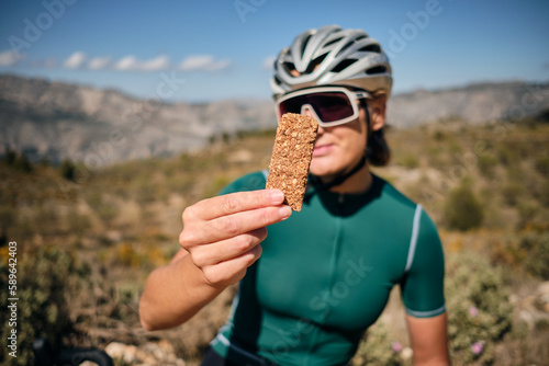 Female cyclist wearing cycling kit holding energy snack for endurance during bicycle training.Sport nutrition.Sport snack for cyclist.Granola energy bar.