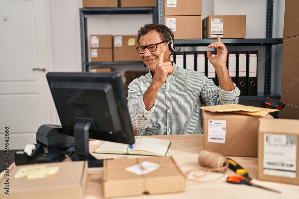 Senior man working at small business ecommerce wearing headset smiling and looking at the camera pointing with two hands and fingers to the side. Senior man working at small business ecommerce wearing headset smiling and looking at the camera pointing with two hands and fingers to the side.