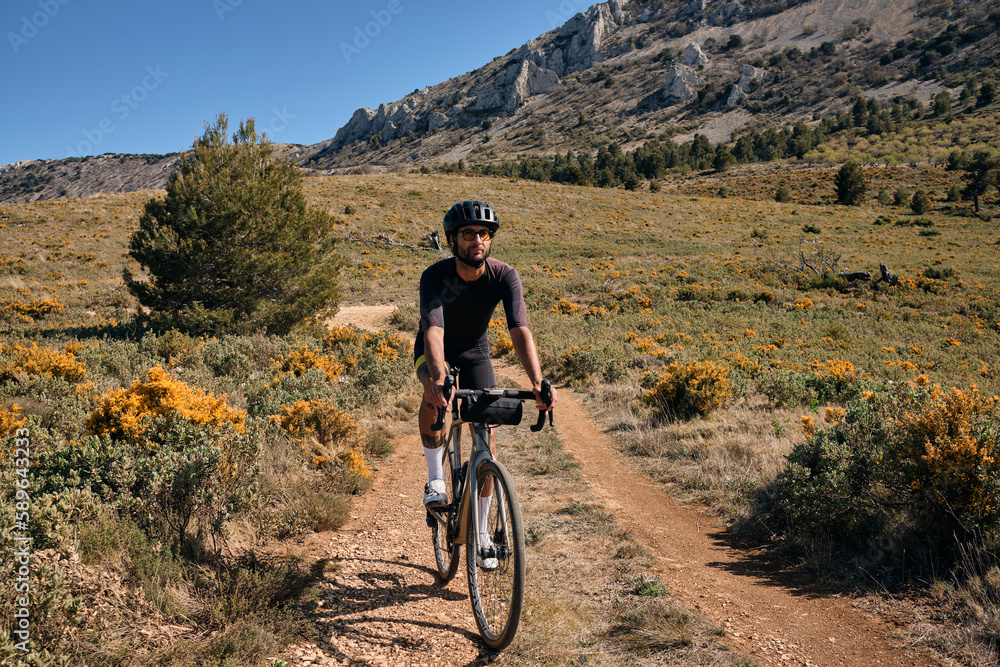 Fototapeta premium Man riding gravel bike on gravel road in mountains with scenic view.Cyclist riding on gravel bike.Cyclist practicing on gravel road.Man cyclist wearing cycling kit and helmet.Alicante region in Spain.