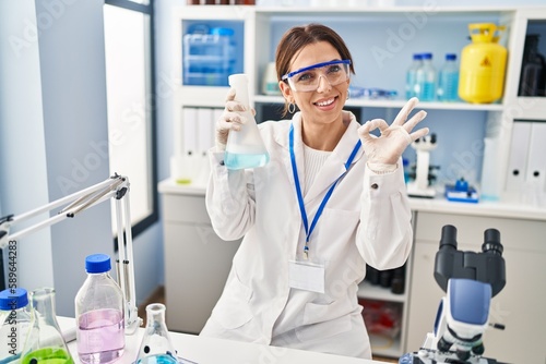 Young brunette woman working at scientist laboratory doing ok sign with fingers, smiling friendly gesturing excellent symbol
