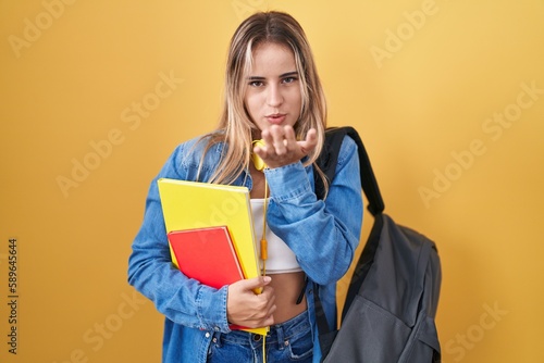 Young blonde woman wearing student backpack and holding books looking at the camera blowing a kiss with hand on air being lovely and sexy. love expression.