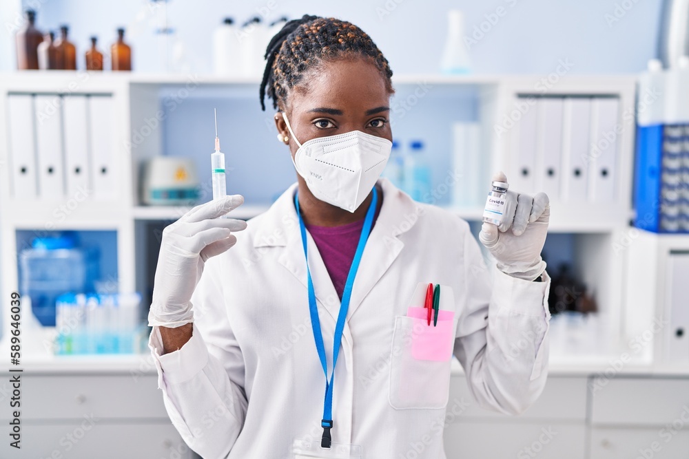African american woman wearing scientist uniform and medical mask holding covid-19 vaccine at laboratory African american woman wearing scientist uniform and medical mask holding covid-19 vaccine at laboratory