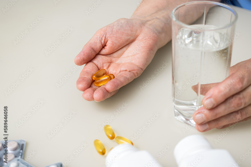Senior woman with wrinkled old hands at the table holding omega 3 yellow capsules, fish oil pills and water glass. Healthcare and medicine concept Senior woman with wrinkled old hands at the table holding omega 3 yellow capsules, fish oil pills and water glass. Healthcare and medicine concept