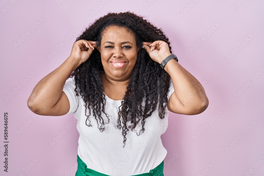 Plus size hispanic woman standing over pink background smiling pulling ears with fingers, funny gesture. audition problem Plus size hispanic woman standing over pink background smiling pulling ears with fingers, funny gesture. audition problem