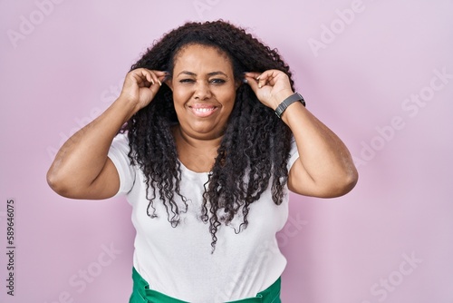 Plus size hispanic woman standing over pink background smiling pulling ears with fingers, funny gesture. audition problem