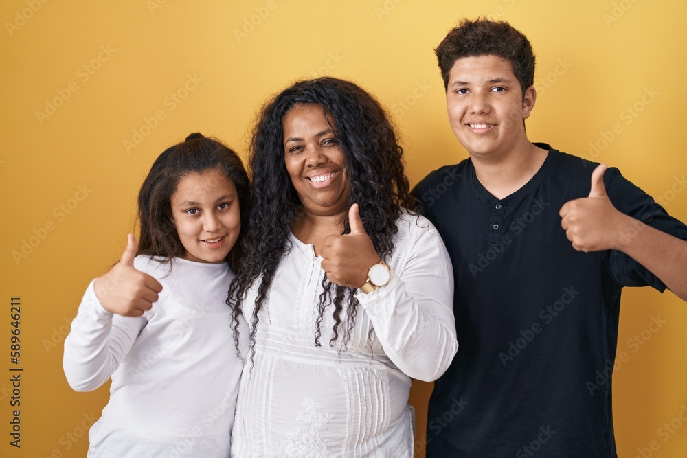 Family of mother, daughter and son standing over yellow background doing happy thumbs up gesture with hand. approving expression looking at the camera showing success. Family of mother, daughter and son standing over yellow background doing happy thumbs up gesture with hand. approving expression looking at the camera showing success.