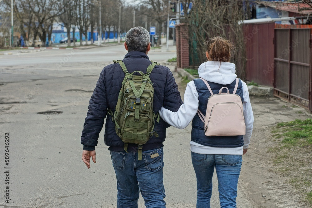 Obraz premium a man and a woman dressed in stylish blue jackets and fashionable blue jeans with comfortable backpacks walk along the asphalt road on an empty street in the spring afternoon