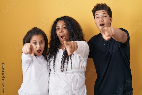 Family of mother, daughter and son standing over yellow background pointing with finger surprised ahead, open mouth amazed expression, something on the front