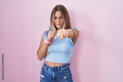 Young blonde woman standing over pink background punching fist to fight, aggressive and angry attack, threat and violence