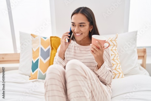 Young beautiful hispanic woman talking on smartphone drinking coffee at bedroom