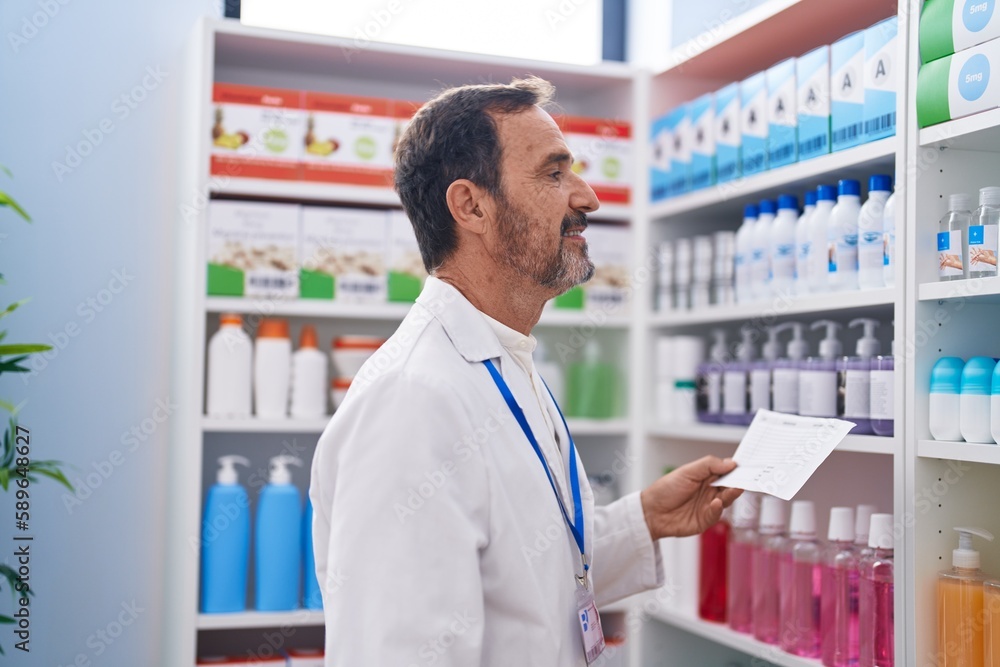 Middle age man pharmacist holding prescription looking to shelving at pharmacy