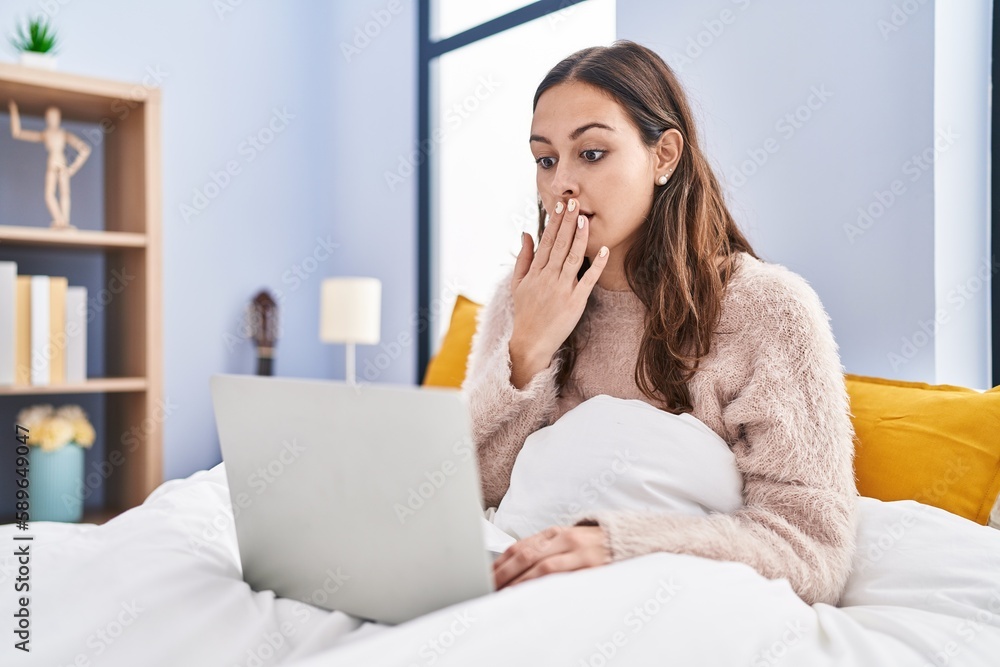 Young hispanic woman using computer laptop on the bed covering mouth with hand, shocked and afraid for mistake. surprised expression Young hispanic woman using computer laptop on the bed covering mouth with hand, shocked and afraid for mistake. surprised expression