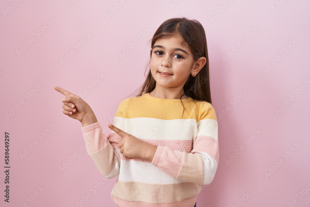 Little hispanic girl wearing sweater over pink background smiling and looking at the camera pointing with two hands and fingers to the side. Little hispanic girl wearing sweater over pink background smiling and looking at the camera pointing with two hands and fingers to the side.