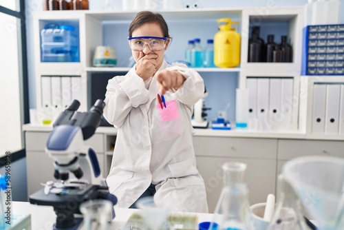 Hispanic girl with down syndrome working at scientist laboratory laughing at you, pointing finger to the camera with hand over mouth, shame expression