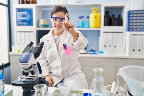 Hispanic girl with down syndrome working at scientist laboratory pointing with finger up and angry expression, showing no gesture