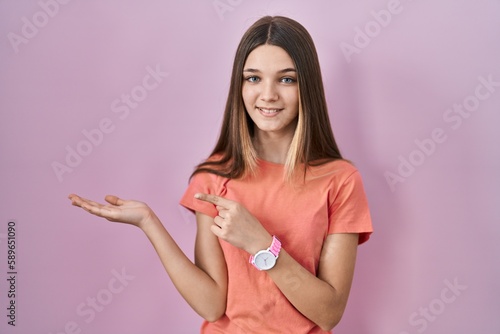 Teenager girl standing over pink background amazed and smiling to the camera while presenting with hand and pointing with finger.
