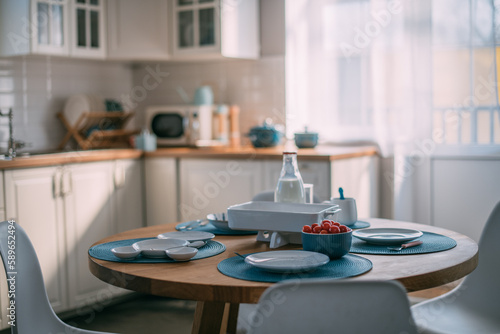 Bright, cozy kitchen in white and blue tones in the house on a sunny day. No one. A table set for dinner.