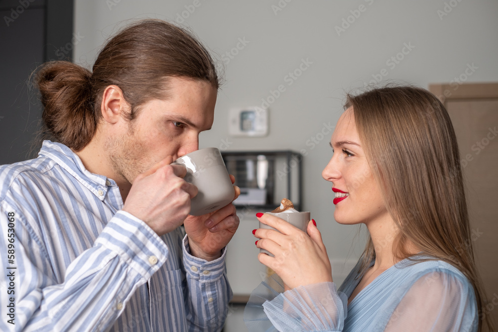 Two young people with beautiful long hair enjoy coffee. The man is thoughtful, the woman watches him with a smile. The shelf is in the background. Copy space Two young people with beautiful long hair enjoy coffee. The man is thoughtful, the woman watches him with a smile. The shelf is in the background. Copy space