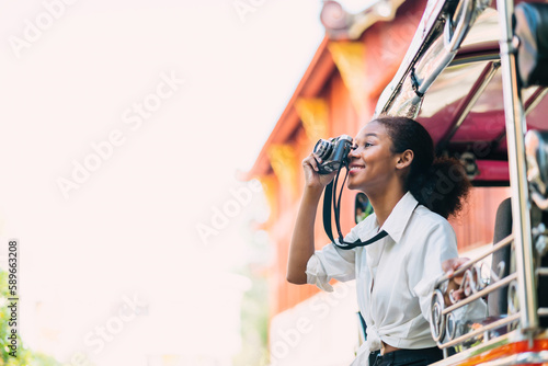 smiling black tourist girl taking photograph outside tuk tuk ride. african american girl using vintage film ride tuk tuk. solo teenage traveler taking picture by old camera enjoying air flow on tuk