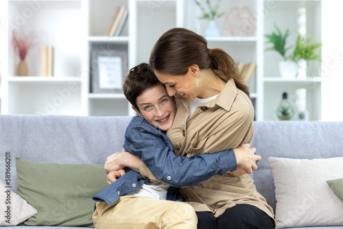 Portrait of a family, mother and teenage son sitting at home on the couch and hugging, having fun, laughing. © Liubomir