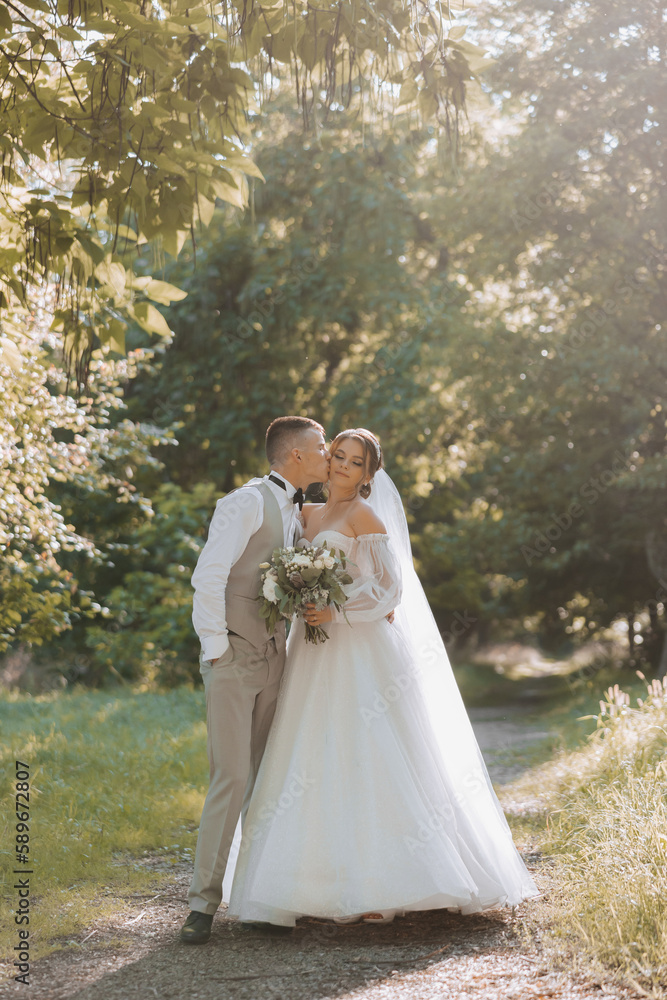 Portrait of a beautiful couple in love on your wedding day. A walk in the park in the sunshine and greenery. Amazing kisses and hugs of the bride and groom with a bouquet