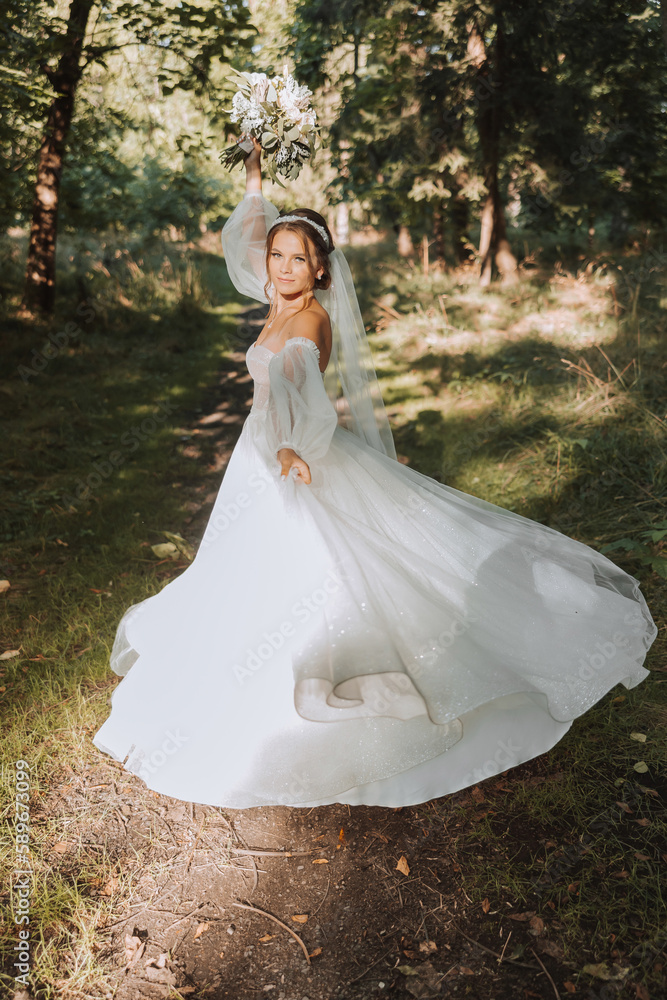 A beautiful bride in a white dress with a long veil is circling with a bouquet in the park. Photo from the back. Young woman, art photo, wedding, bride, open shoulders. light.